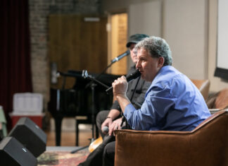 Music producer’s album pays homage to Mercer heritage, American heroes Man in a blue shirt speaks into a microphone while seated on a stage during an indoor event.