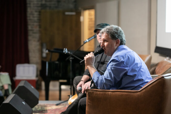 Man in a blue shirt speaks into a microphone while seated on a stage during an indoor event.