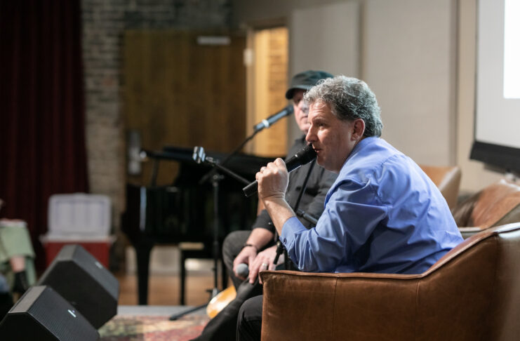 Man in a blue shirt speaks into a microphone while seated on a stage during an indoor event.
