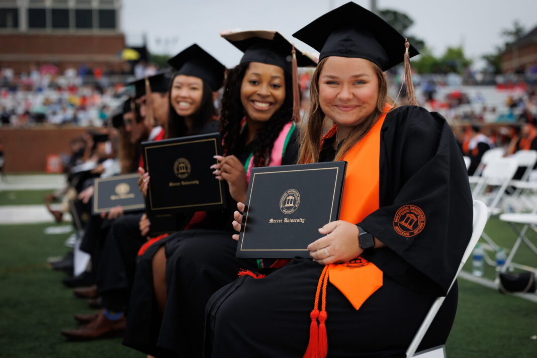 Mercer graduates in caps and gowns sit in rows holding diplomas during an outdoor graduation ceremony.