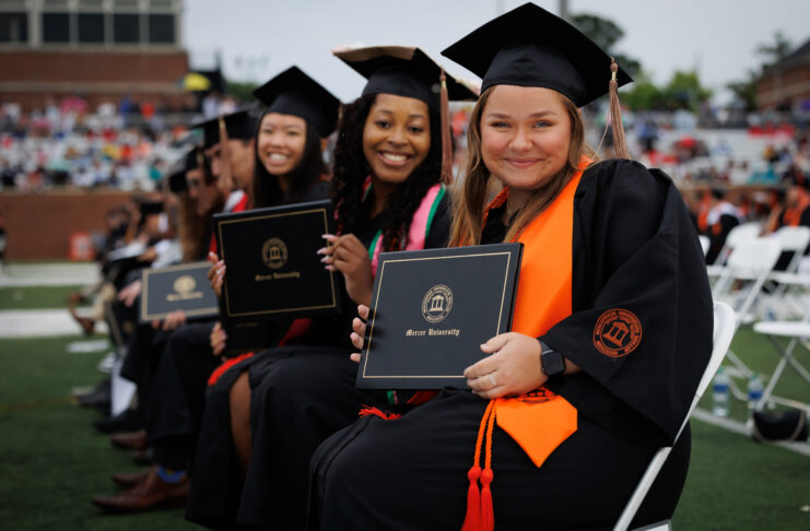 Mercer graduates in caps and gowns sit in rows holding diplomas during an outdoor graduation ceremony.