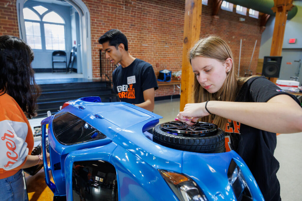 Students work together on assembling a blue toy car indoors, focusing on attaching its wheels.