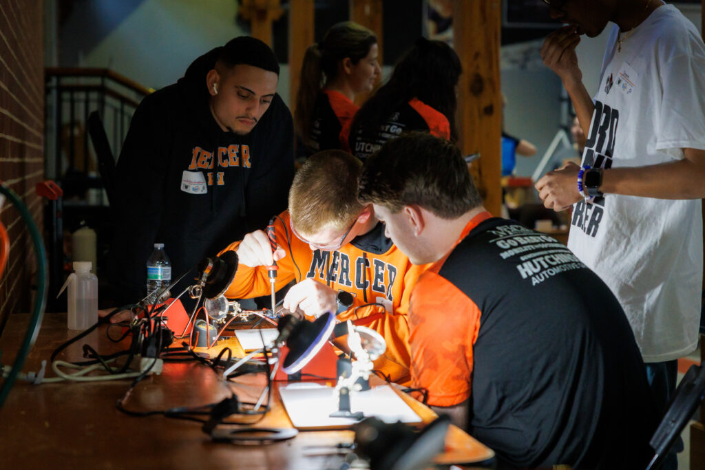 Three students in orange Mercer shirts work on an electronics project at a table while others observe nearby.