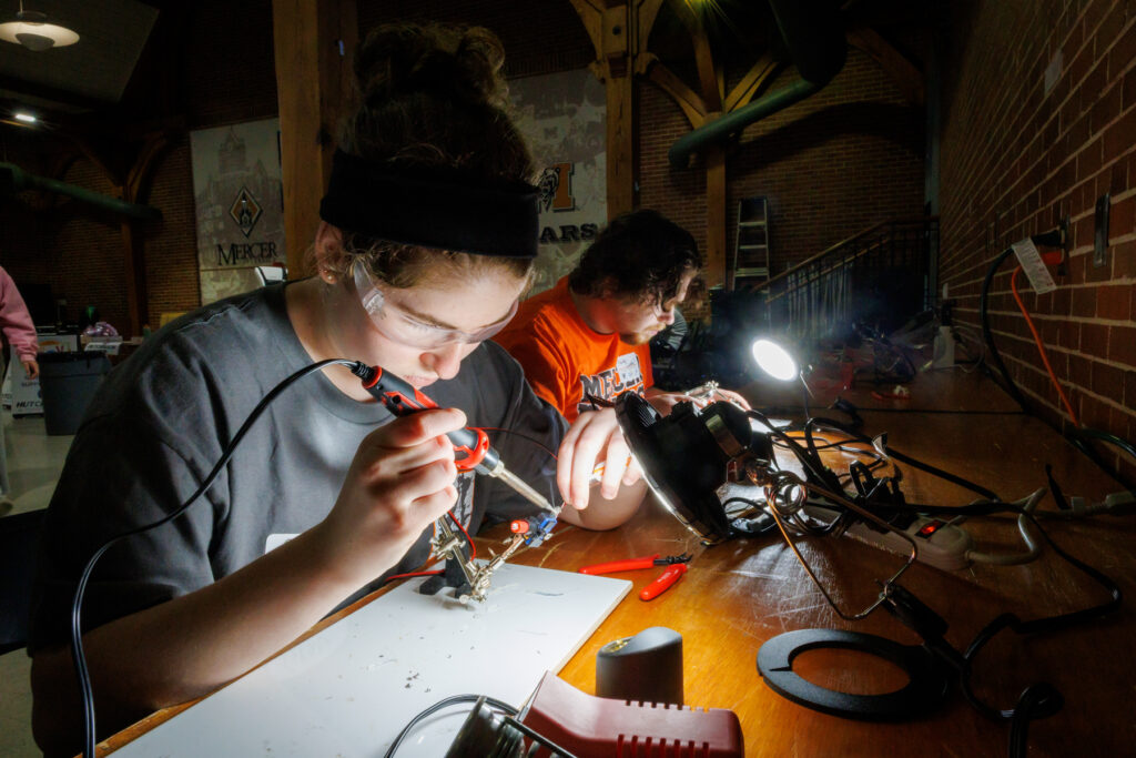 Two people wearing safety glasses solder electronic components at a workbench under bright task lights.