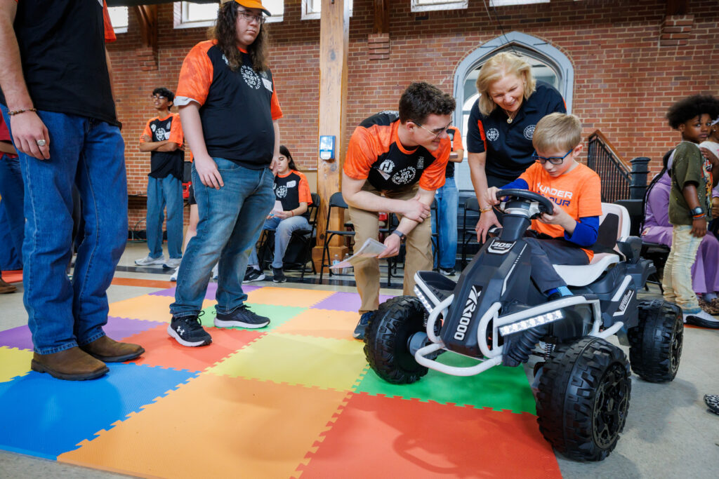 A child in a toy car is guided by adults on a colorful mat at an indoor event, with other people observing.