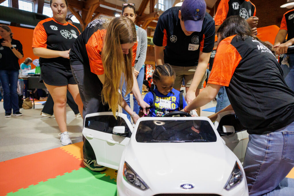 A child is helped into a small white toy car by Mercer students at Go Baby Go.