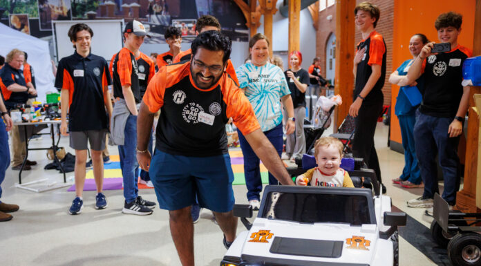 Mercer’s Go Baby Go changes kids’ lives one vehicle at a time A Mercer student guides a small child riding a toy car, surrounded by smiling people in Penfield Hall.