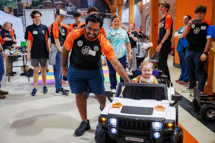 A Mercer student guides a small child riding a toy car, surrounded by smiling people in Penfield Hall.