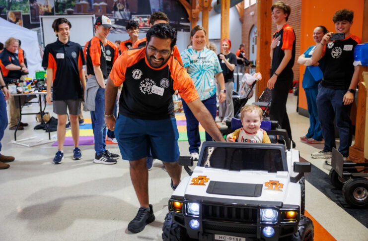A Mercer student guides a small child riding a toy car, surrounded by smiling people in Penfield Hall.