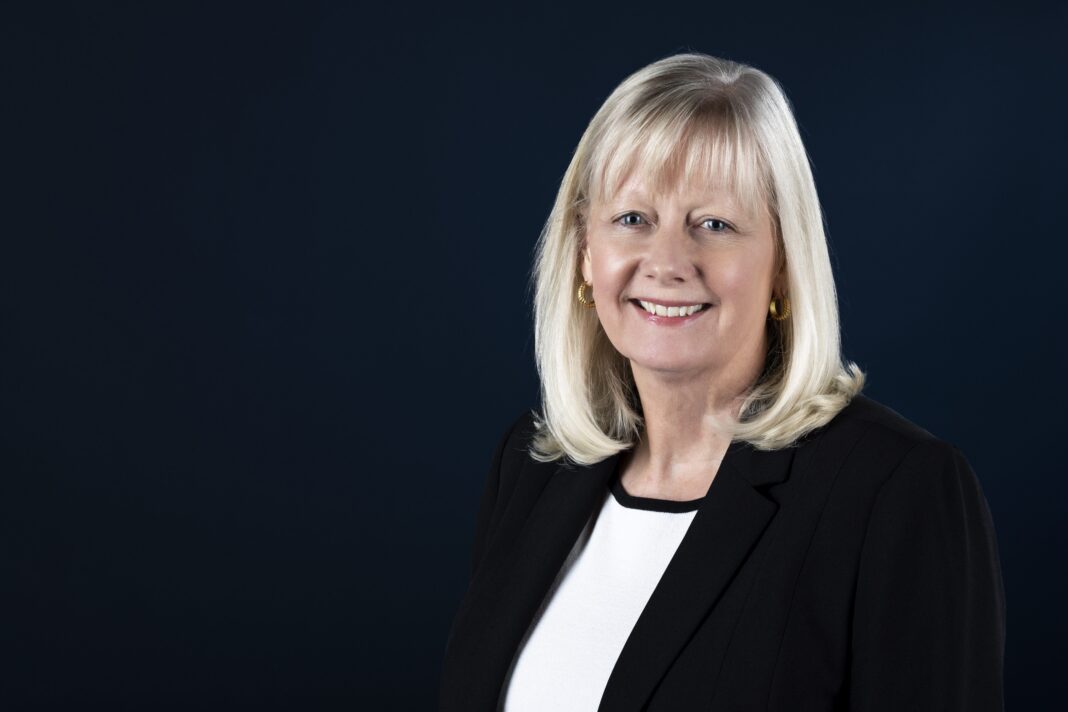 Woman with shoulder-length blonde hair in a black blazer and white blouse, smiling against a dark blue background.
