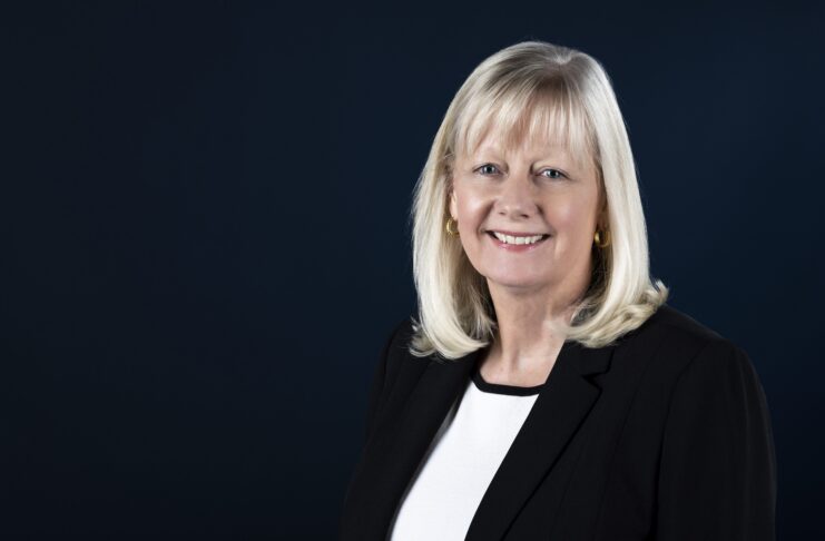 Woman with shoulder-length blonde hair in a black blazer and white blouse, smiling against a dark blue background.