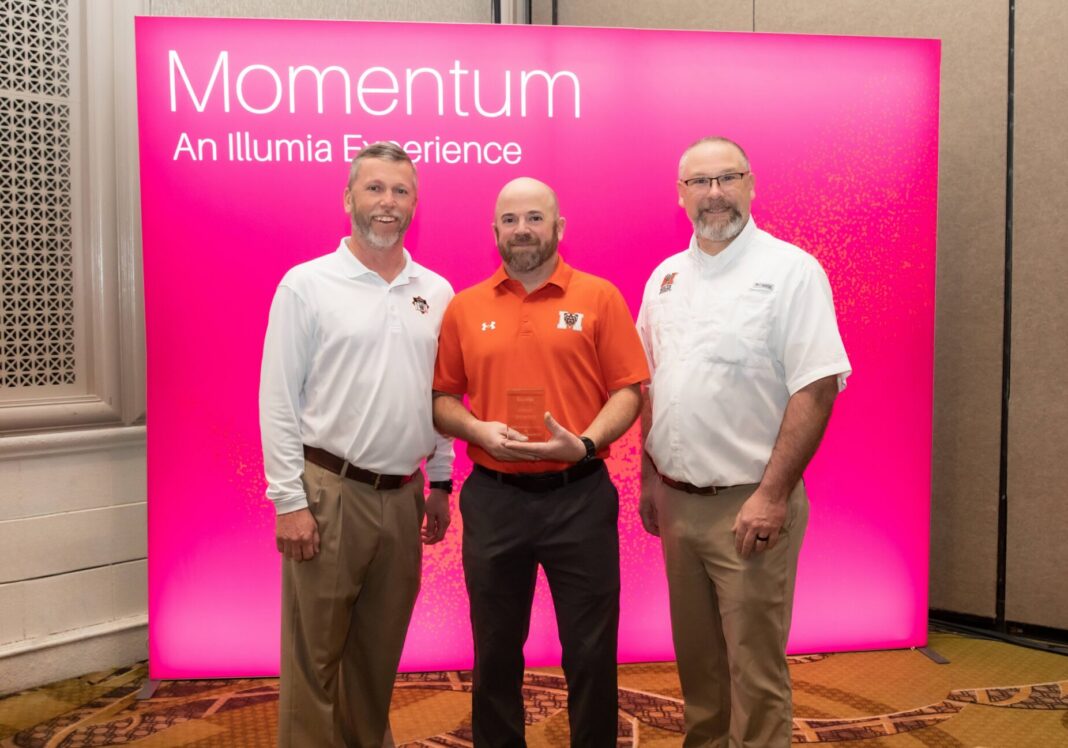 Three men stand in front of a pink Momentum: An Illumia Experience sign, with one holding a clear award.