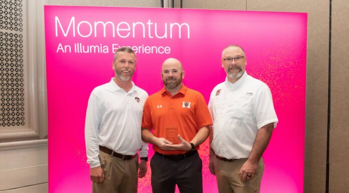 Auxiliary Services receives Illumia Distinction Award for Innovation Excellence Three men stand in front of a pink Momentum: An Illumia Experience sign, with one holding a clear award.