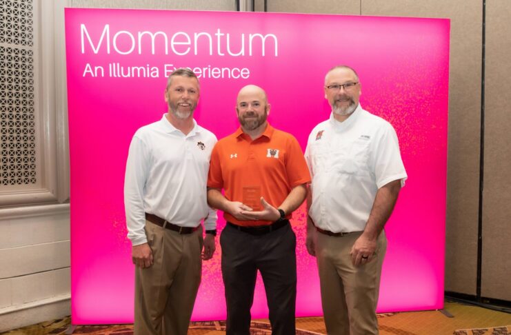 Three men stand in front of a pink Momentum: An Illumia Experience sign, with one holding a clear award.
