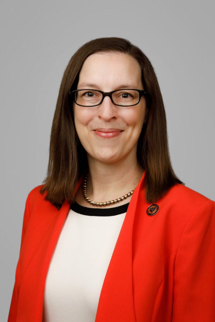 Woman with straight brown hair wearing glasses, a red blazer, white top, and a beaded necklace, smiling at the camera.