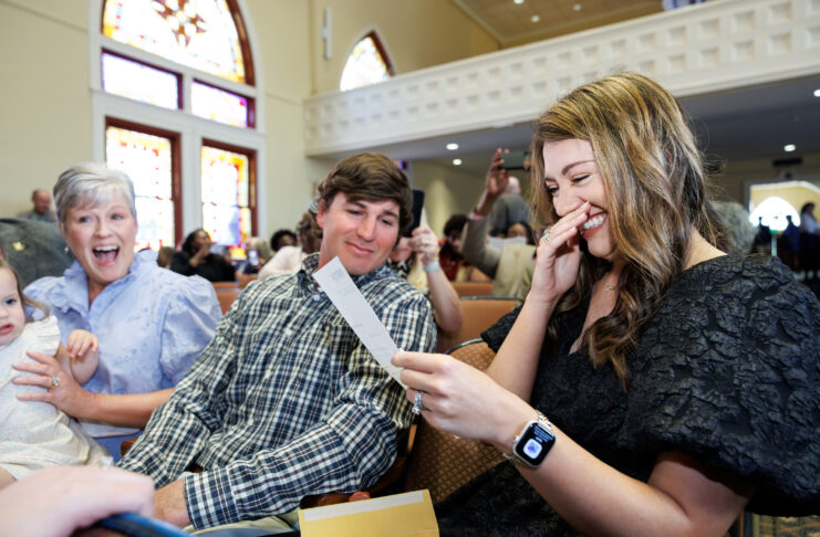 A young woman smiles and covers her mouth while reading a card, seated with two others in a church.