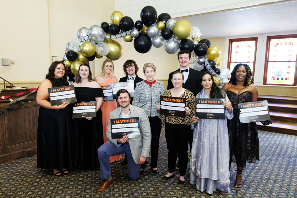A group of people in formal attire pose indoors with I Matched! signs in front of a balloon arch.