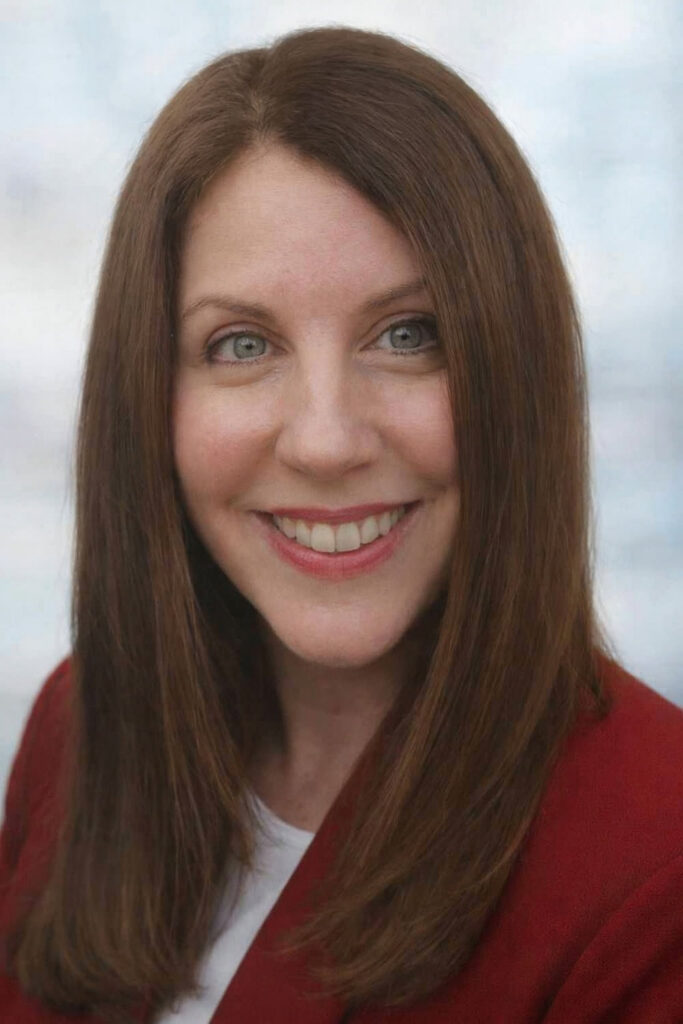 Woman with straight brown hair, smiling, wearing a red blazer and white top, posing in front of a light background.