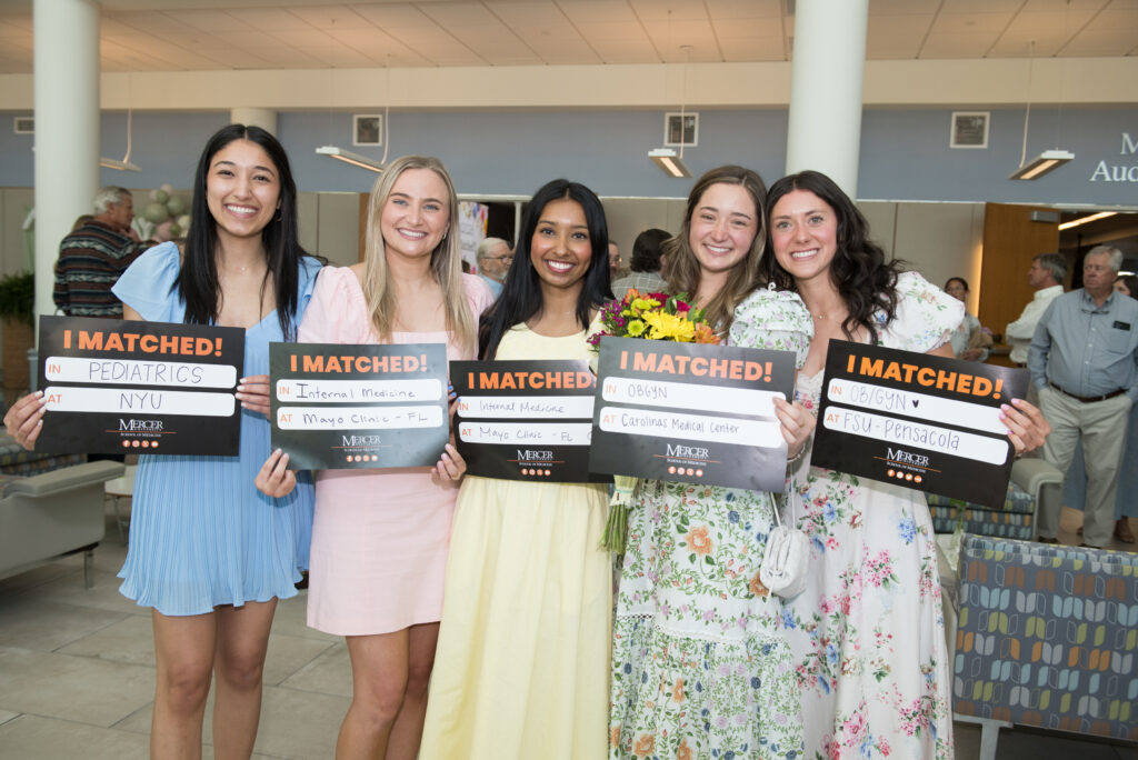 Five women stand indoors holding signs announcing their medical residency match results, smiling at the camera.