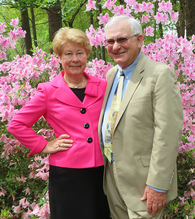 An older woman and man stand together in front of blooming pink flowers, both smiling and wearing formal attire.