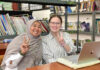 Mercer alumna serves as Peace Corps volunteer in Indonesia Two women sit at a table in a library, smiling and making peace signs, with a laptop and books in the background.