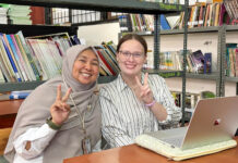 Mercer alumna serves as Peace Corps volunteer in Indonesia Two women sit at a table in a library, smiling and making peace signs, with a laptop and books in the background.
