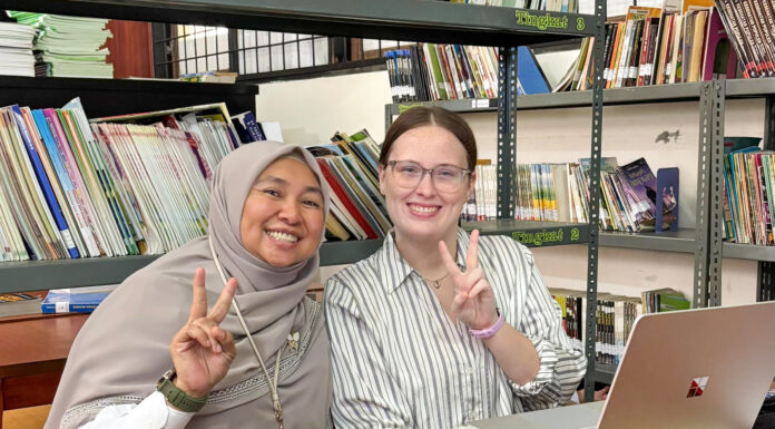 Mercer alumna serves as Peace Corps volunteer in Indonesia Two women sit at a table in a library, smiling and making peace signs, with a laptop and books in the background.