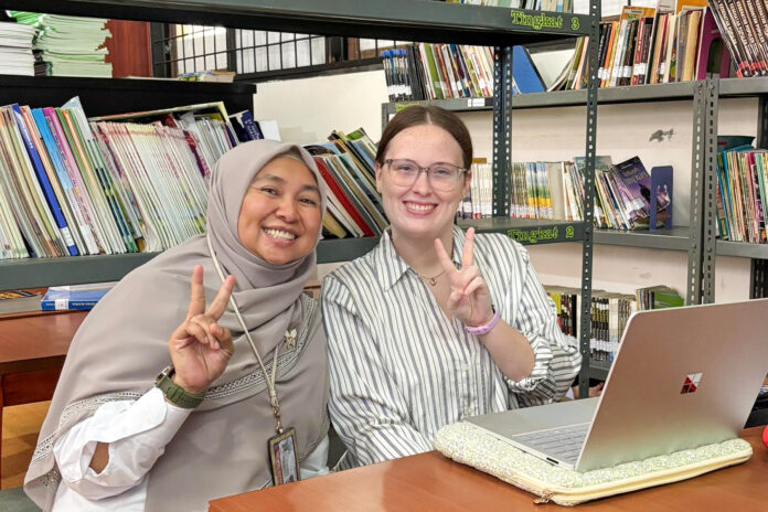 Two women sit at a table in a library, smiling and making peace signs, with a laptop and books in the background.