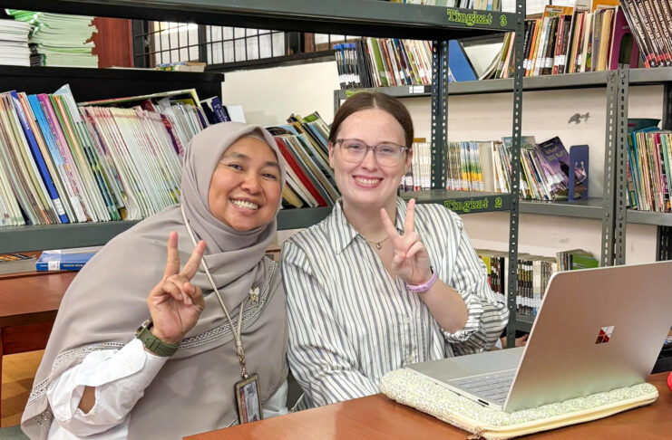 Two women sit at a table in a library, smiling and making peace signs, with a laptop and books in the background.