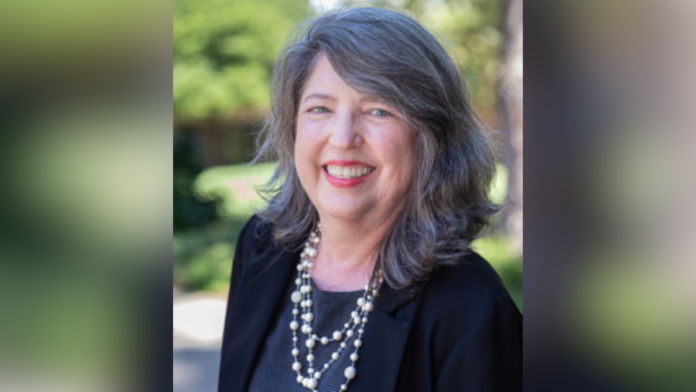 Middle-aged woman with long gray hair, wearing a black blazer and pearl necklace, smiling outdoors.
