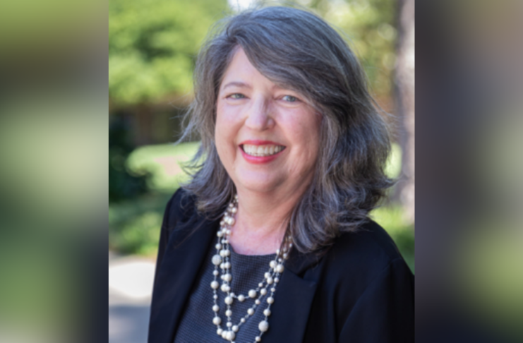 Middle-aged woman with long gray hair, wearing a black blazer and pearl necklace, smiling outdoors.
