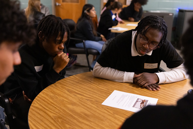 Three students sit at a round table, reading and discussing a printed document in a classroom setting.