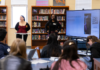 Mercer students introduce high school juniors, seniors to anthropology Two presenters stand at the front of a library classroom, speaking to students with a large screen displaying a question and QR code.
