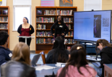 Mercer students introduce high school juniors, seniors to anthropology Two presenters stand at the front of a library classroom, speaking to students with a large screen displaying a question and QR code.