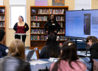 Mercer students introduce high school juniors, seniors to anthropology Two presenters stand at the front of a library classroom, speaking to students with a large screen displaying a question and QR code.