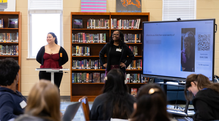 Mercer students introduce high school juniors, seniors to anthropology Two presenters stand at the front of a library classroom, speaking to students with a large screen displaying a question and QR code.