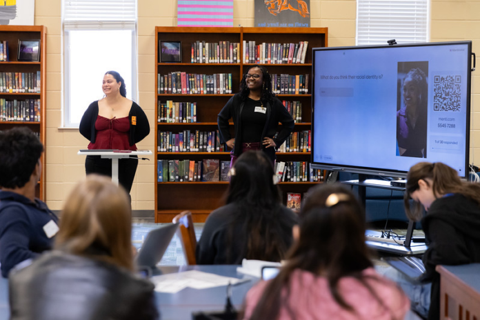 Two presenters stand at the front of a library classroom, speaking to students with a large screen displaying a question and QR code.