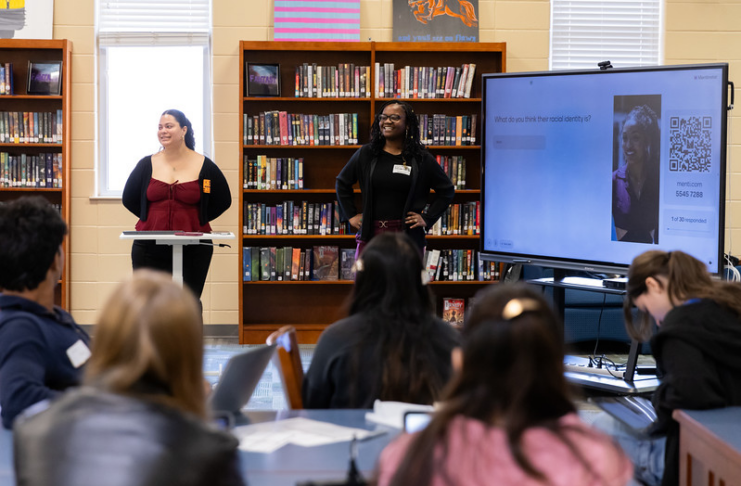 Two presenters stand at the front of a library classroom, speaking to students with a large screen displaying a question and QR code.