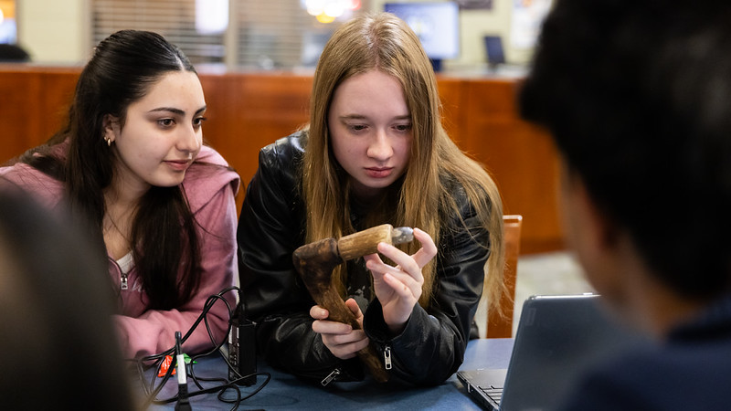 Two students examine a wooden object closely while sitting at a table with electronic equipment and a laptop.