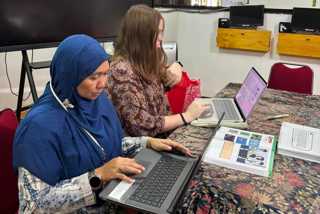 Two women work on laptops at a table with workbooks in a classroom setting.