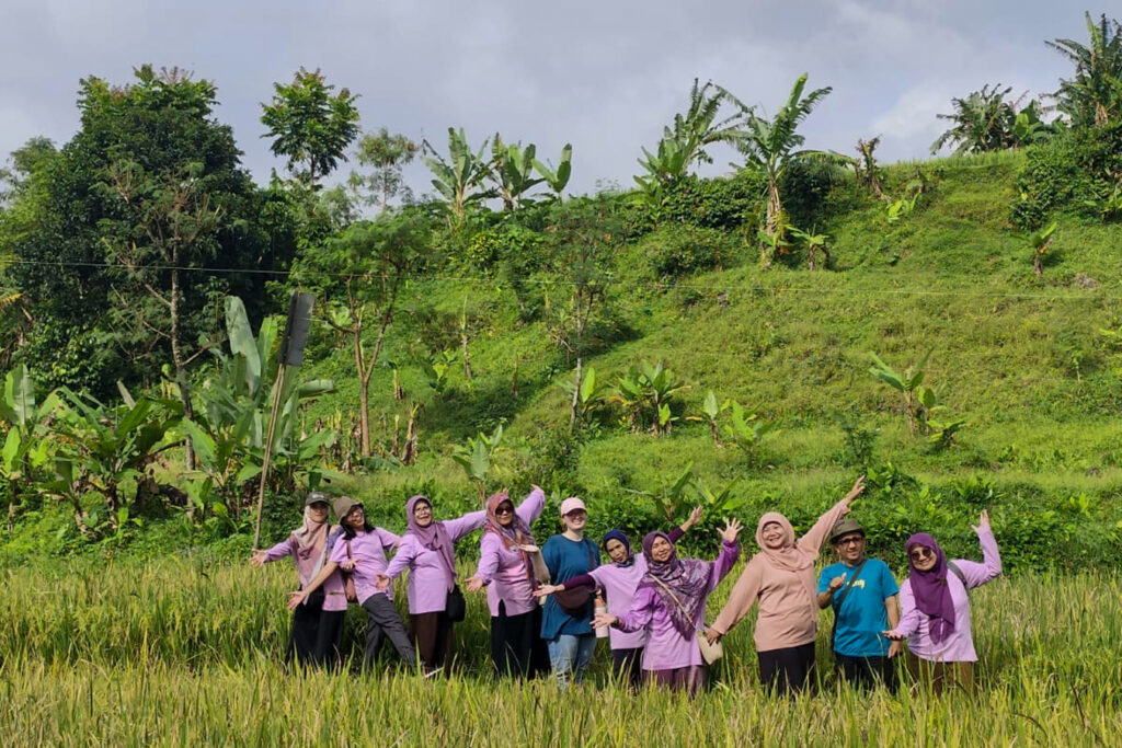 A group of people in purple shirts standing and posing in a green field with trees and a grassy hill in the background.