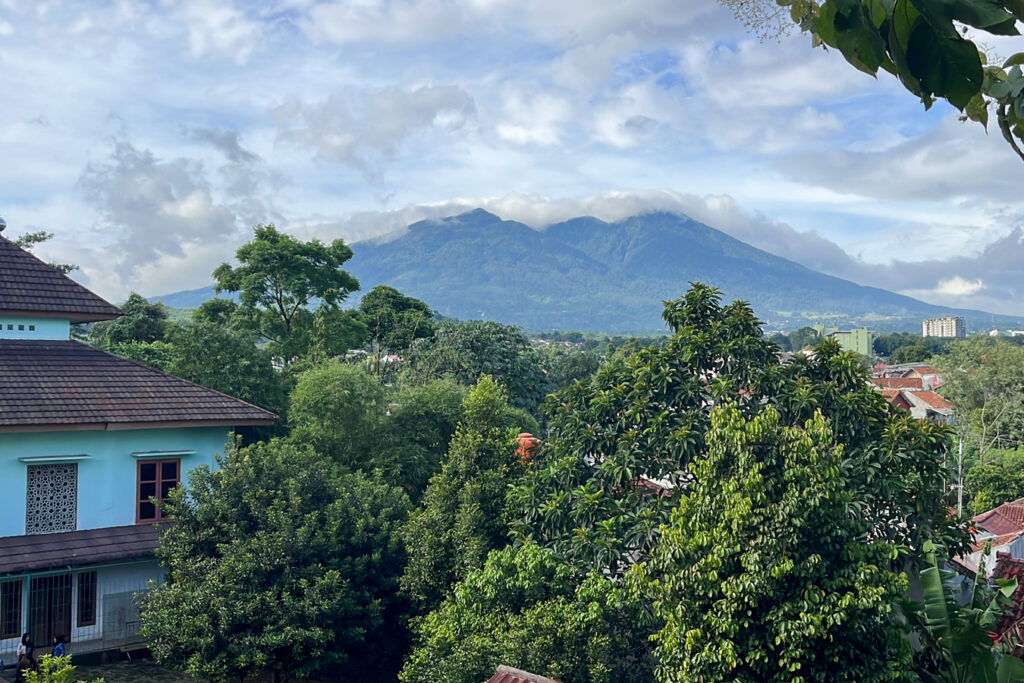 A lush green landscape with trees and buildings in front of a large, cloud-covered mountain under a partly cloudy sky.