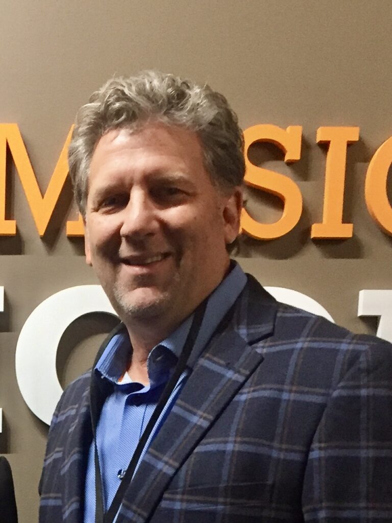 Man with gray hair in a plaid blazer and blue shirt standing in front of a wall with large orange and white letters.