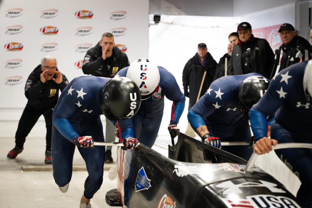 Four USA bobsledders start a race as coaches and officials look on in an indoor track setting.