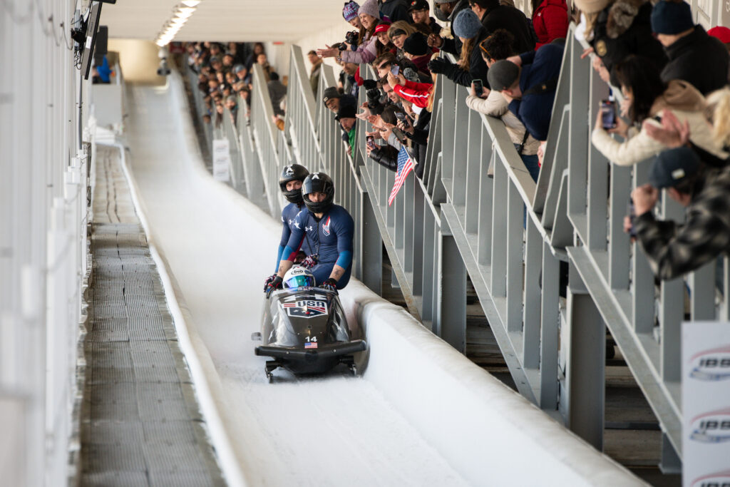 A four-person bobsled team races down an icy track as spectators watch and cheer from the stands above.