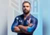Mercerian competes in bobsled events in Winter Olympics A male athlete in a blue USA uniform stands with arms crossed in front of a blurred USA logo background.