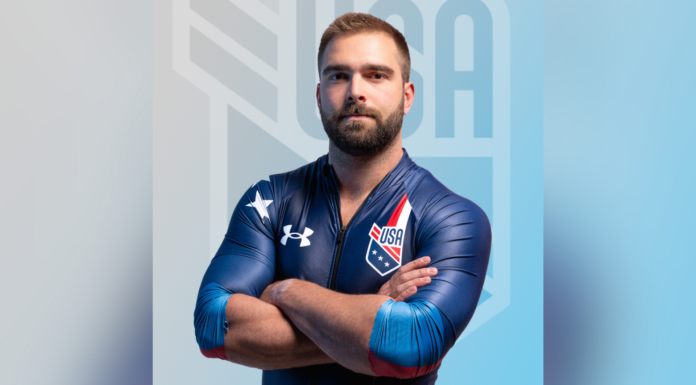 Mercerian competes in bobsled events in Winter Olympics A male athlete in a blue USA uniform stands with arms crossed in front of a blurred USA logo background.