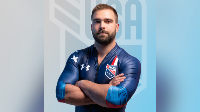 A male athlete in a blue USA uniform stands with arms crossed in front of a blurred USA logo background.