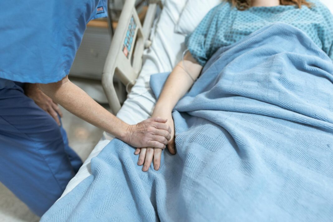 A health care worker holds the hand of a patient lying in a hospital bed covered with a blue blanket.