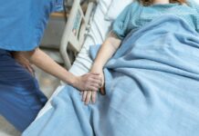 Mercer professors awarded grant to explore how to better support dying patients A healthcare worker holds the hand of a patient lying in a hospital bed covered with a blue blanket.